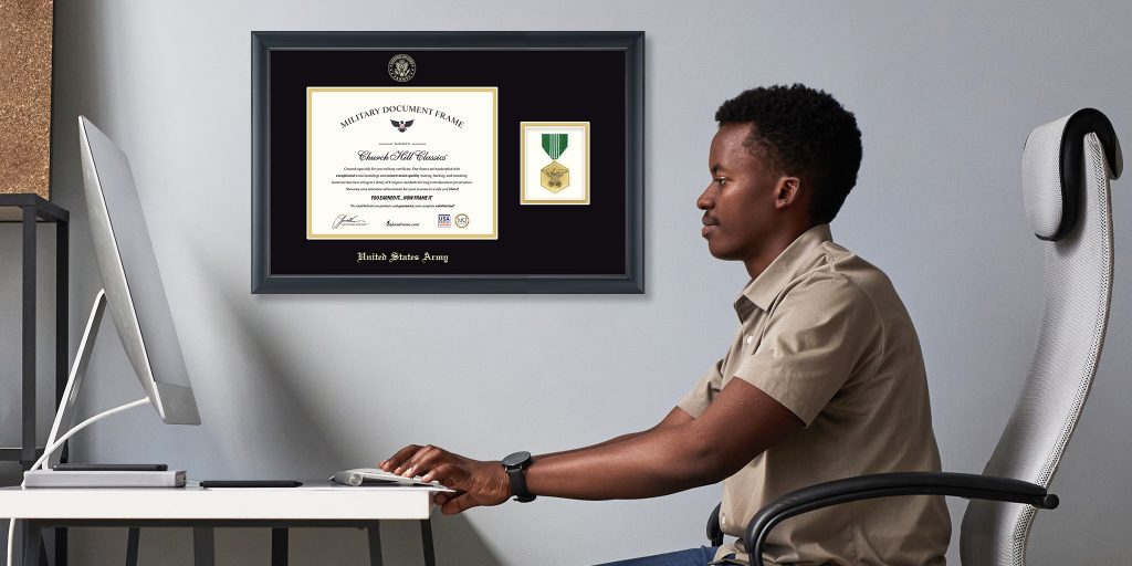 Service member working at a desk with custom military certificate and medal frame hung on the wall