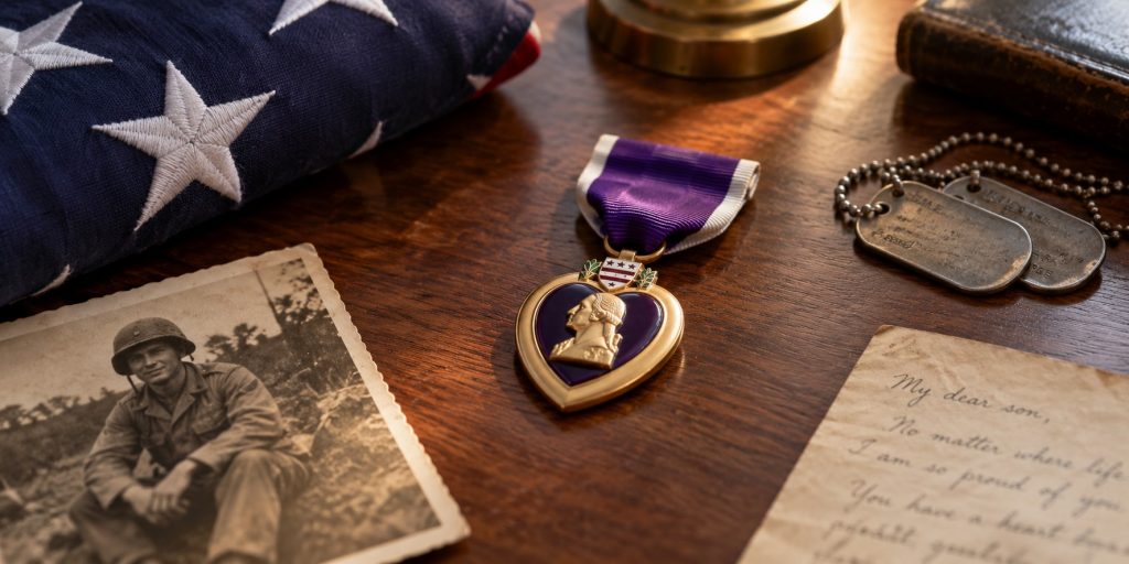 Purple Heart medal displayed on a wooden desk with a folded American flag, dog tags, and a vintage military photo